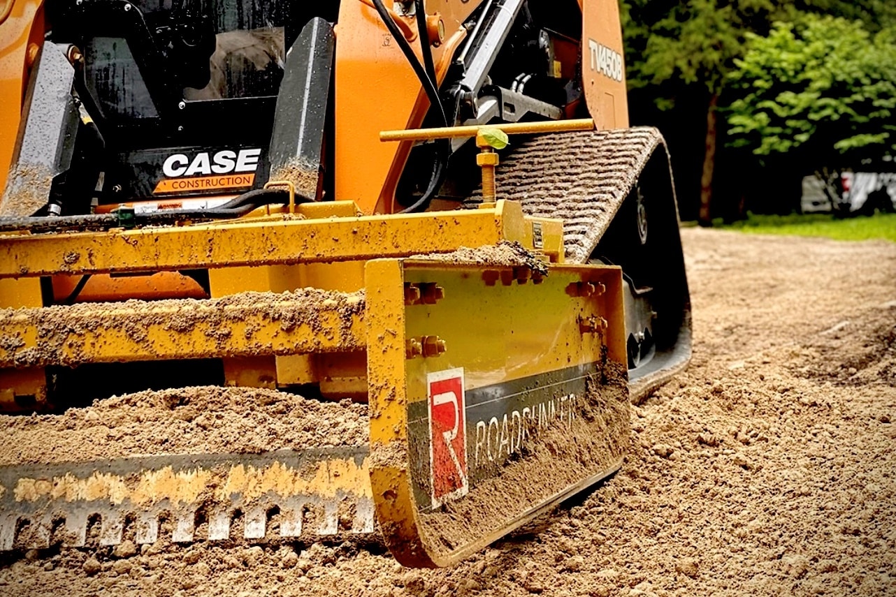 Compact tractor with Roadrunner grader blade smoothing a gravel road, showcasing dual blades, rugged steel frame, and precise grading action